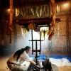 A man, surrounded by fish skins hanging to dry, kneels before a large pit as he demonstrates the tanning process.