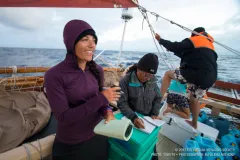 Four people working aboard the deck of a ship. Two are checking materials and noting observations while the two in the background adjust the canoe's rigging.