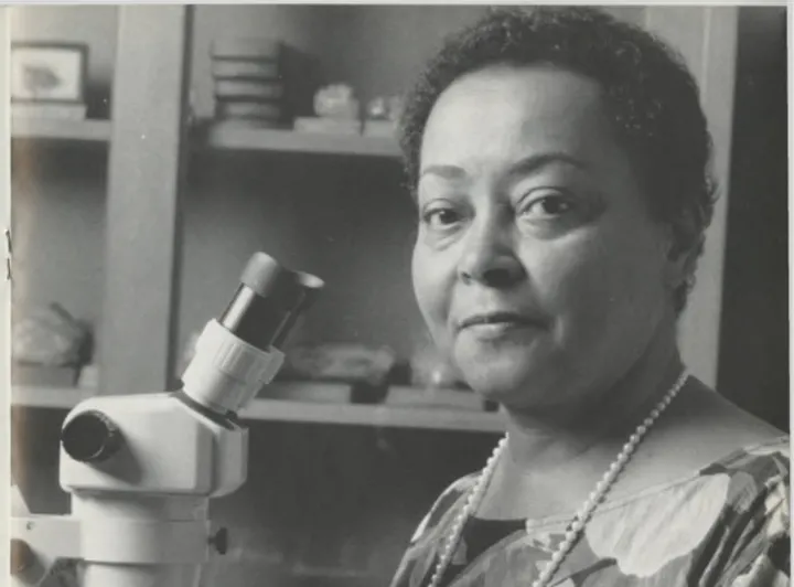 a woman with short black hair and a long necklace of pearls sits in front of a microscope and holds a small button coral